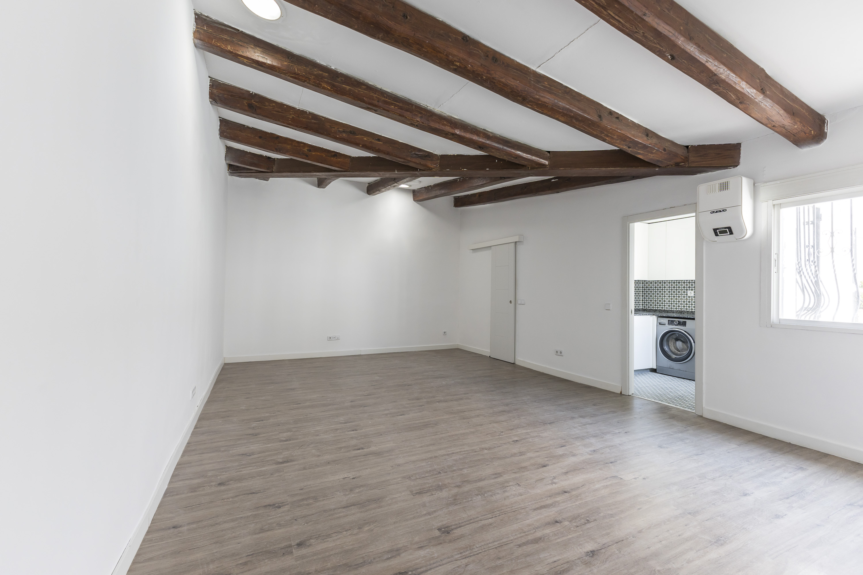 Empty room with white walls, wood-look flooring, and exposed dark wood ceiling beams, with a view into a small laundry area. [AI-generated text]