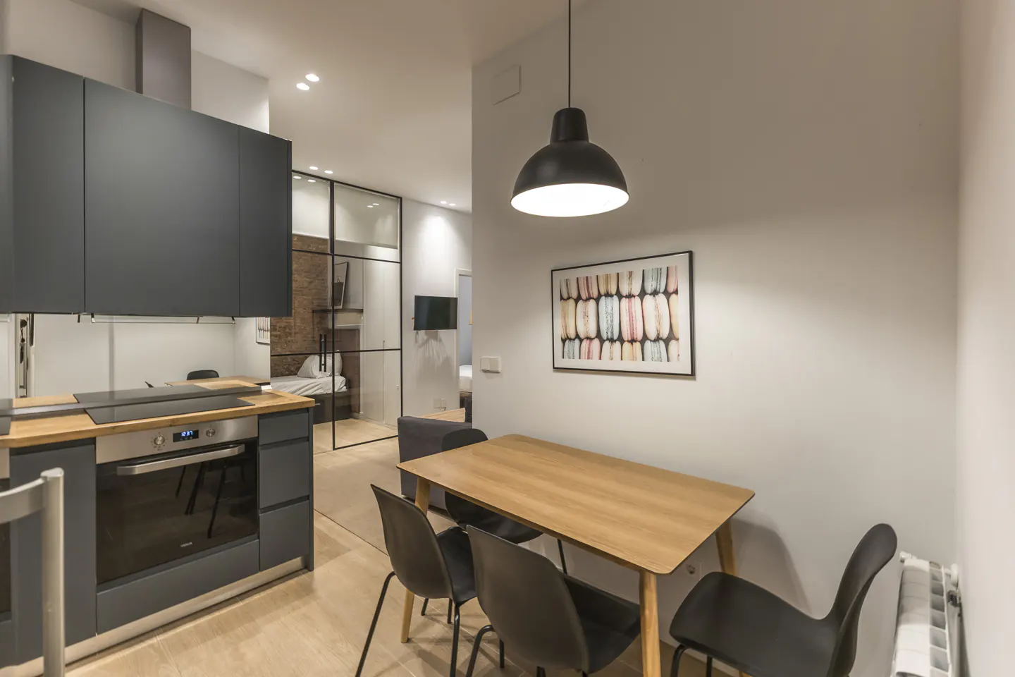 A modern apartment interior shows a kitchen with gray cabinets, a wooden countertop, and a stainless steel oven. A dining area features a wooden table with black chairs under a black pendant light. A glass-enclosed bedroom is visible in the background.