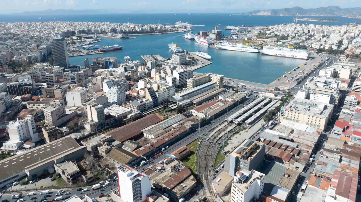 Aerial view of Piraeus port in Greece, featuring multiple large ferries docked in the blue harbor surrounded by city buildings.