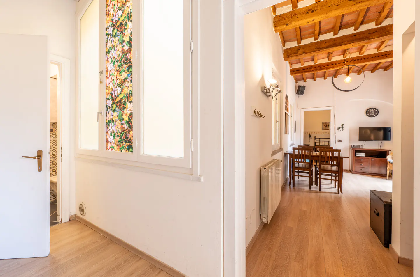 Hallway view of a home interior with wood floors, white walls, and a window with floral film. A dining area is visible in the background.