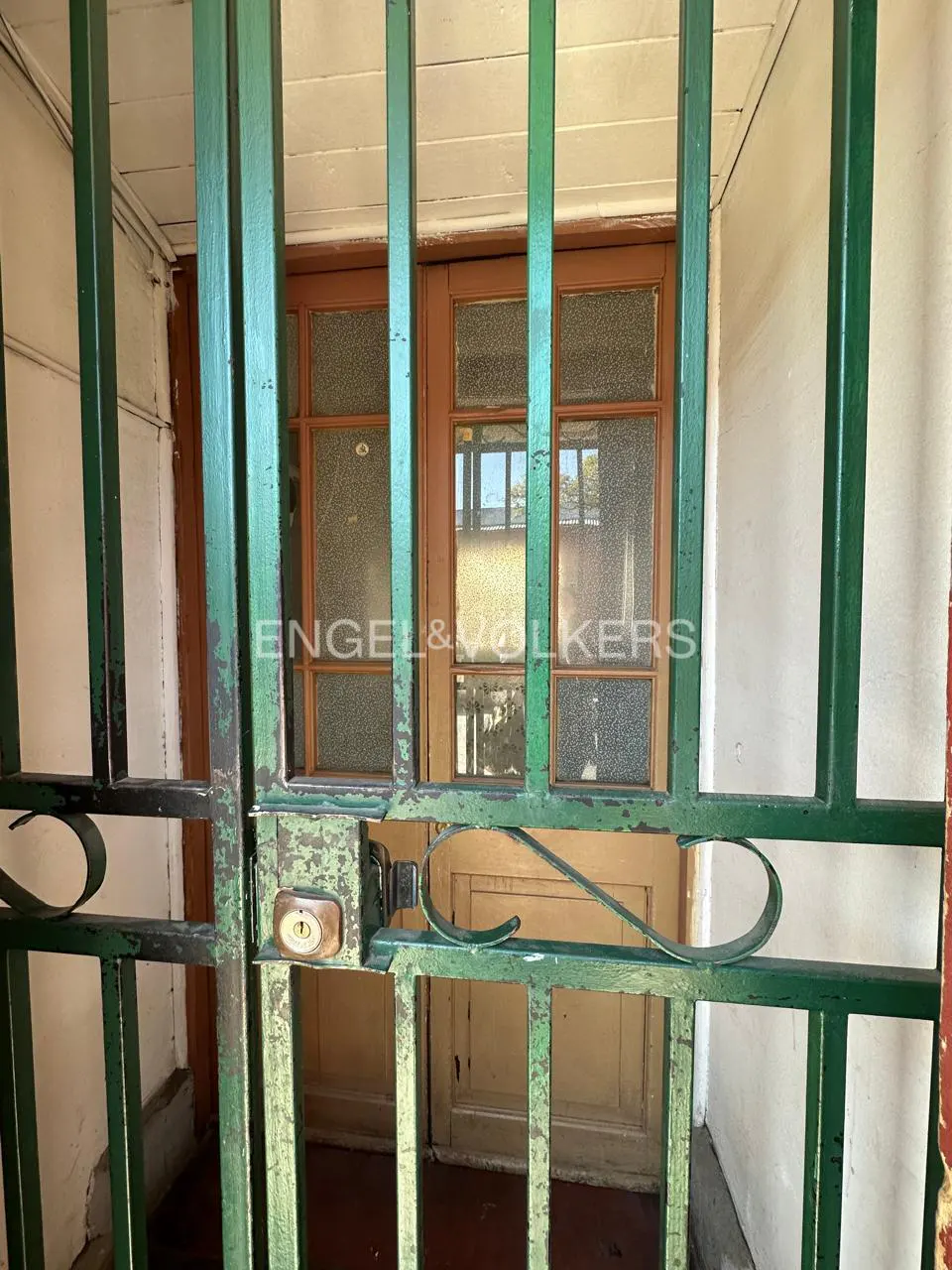 A green metal security gate with a lock stands in front of a wooden door with glass panels at a home entrance.