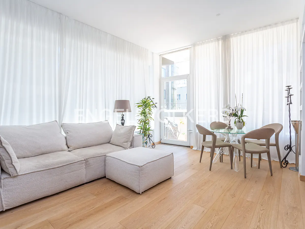 Bright living room with a beige sofa, ottoman, glass table with four chairs, and light wood floors. White curtains cover the windows.