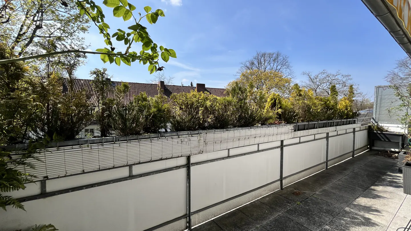 A balcony with a white wall and green bushes, with a view of trees and a blue sky.