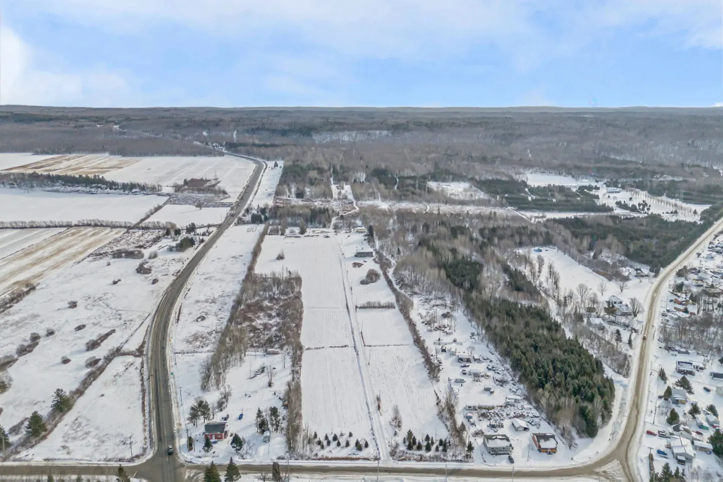 Aerial view of snow-covered fields, forests, and roads under a blue sky. Houses and buildings are scattered throughout the landscape.