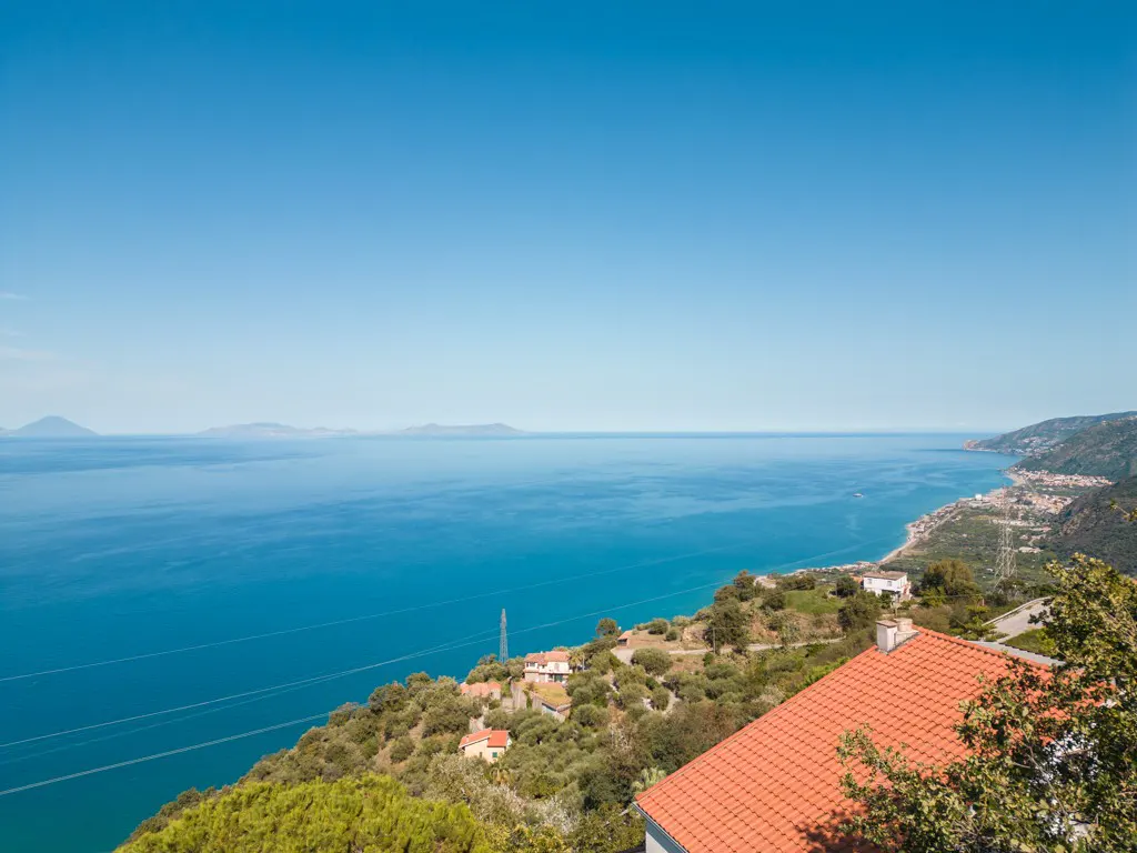 High-angle view of a Mediterranean coastline with a red-tiled roof in the foreground and a vast blue sea under a clear sky.
