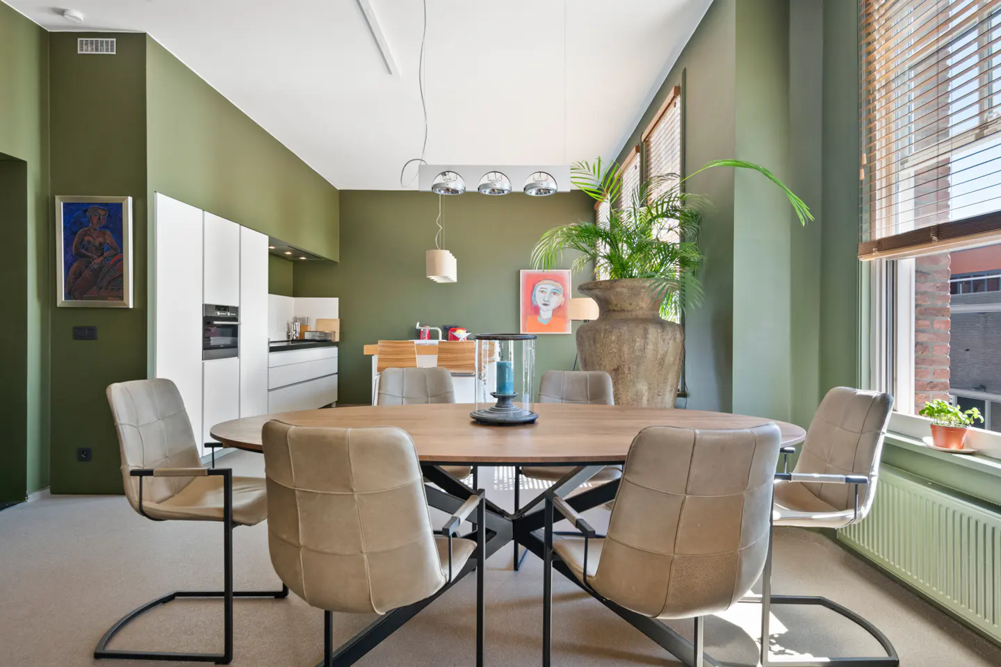 A dining room with olive green walls, a round wooden table, and six beige chairs. A large plant sits near a window with blinds.