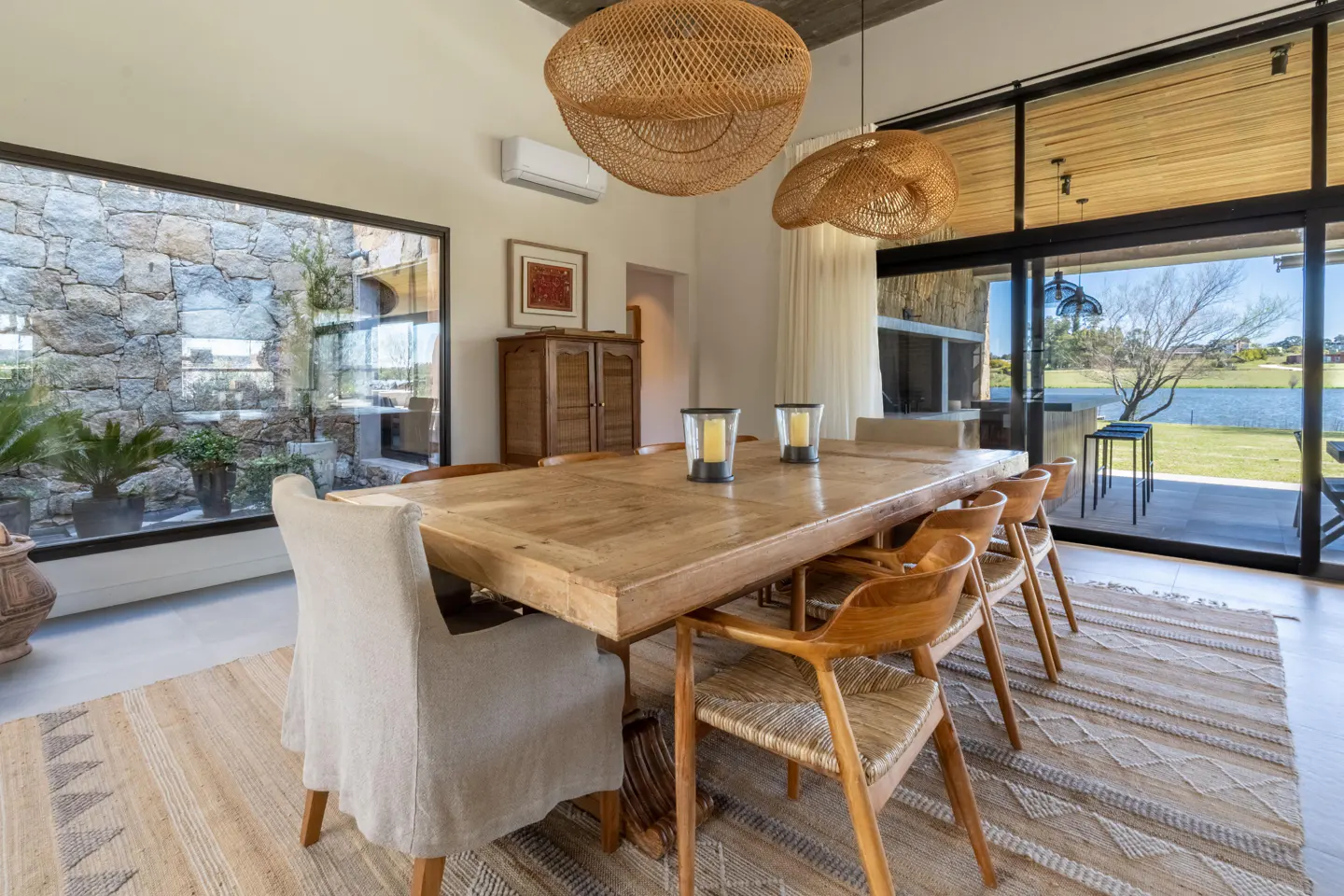 A dining room with a long wooden table, chairs, and woven light fixtures. A large window shows a stone wall and greenery.