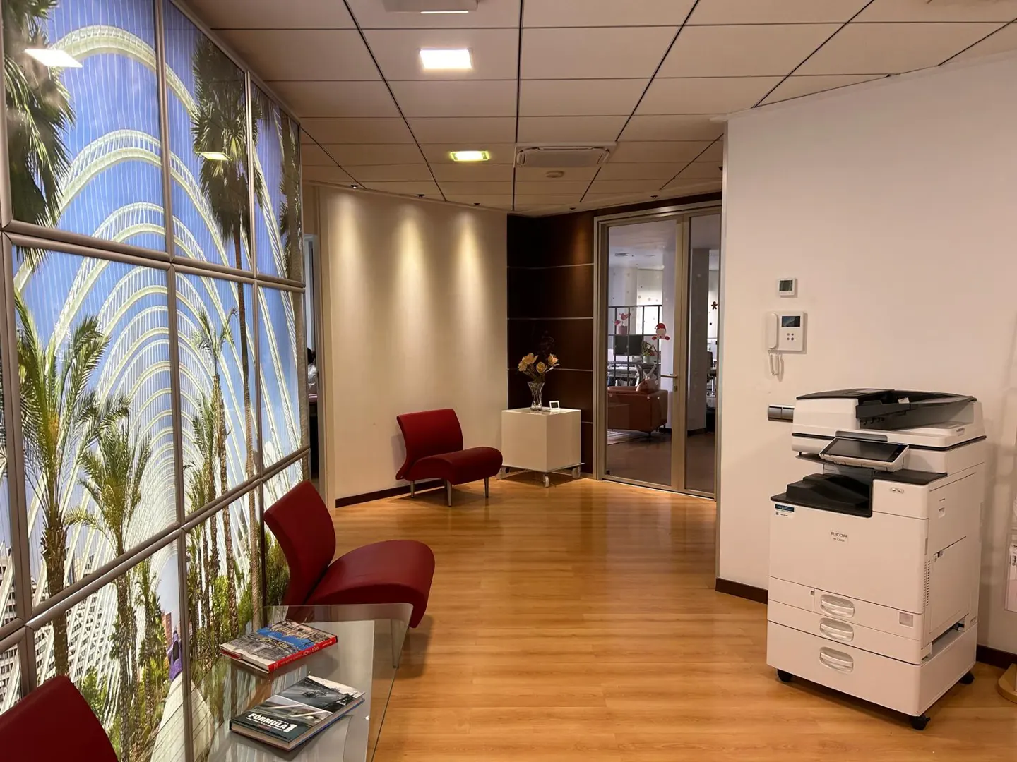 Office waiting area with red chairs, glass table, and a large window with a palm tree image. A printer is on the right.