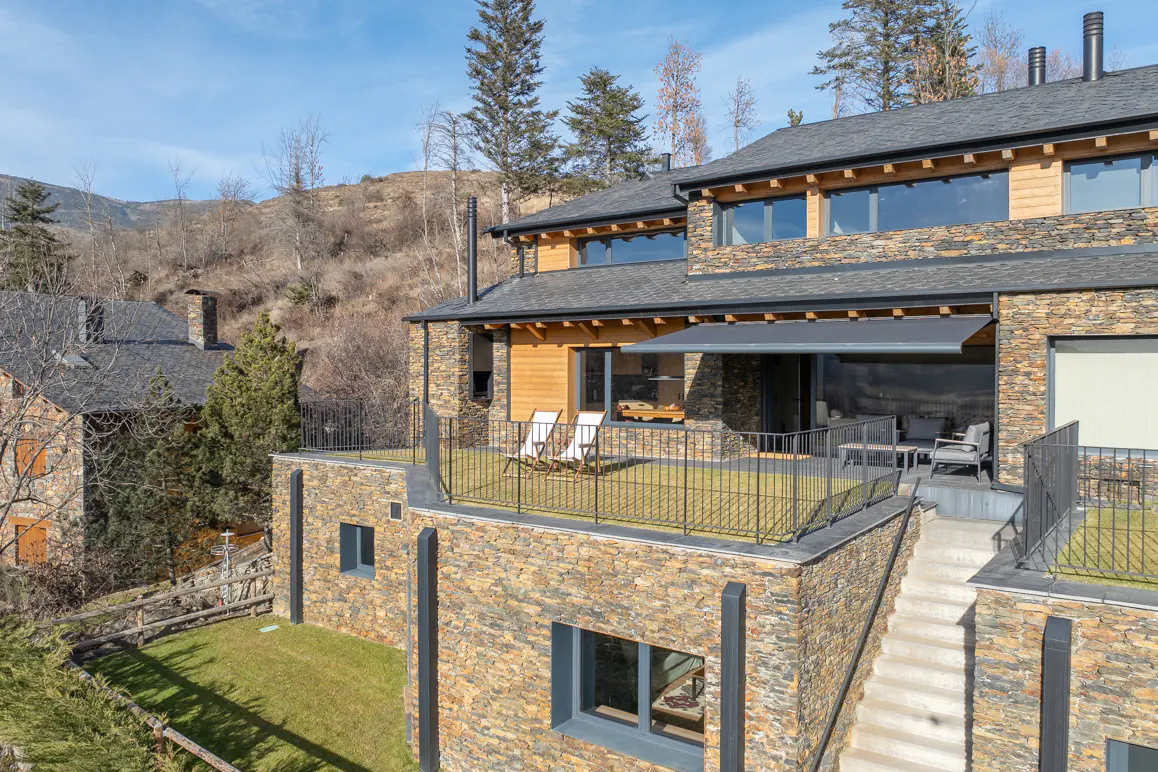 Exterior view of a stone house with a rooftop terrace, chairs, and mountain backdrop.