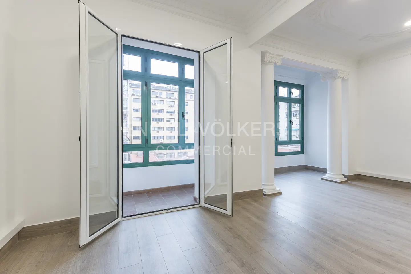 Bright, empty room with light wood floors, white walls, and open glass doors to a balcony with green window frames and city views.