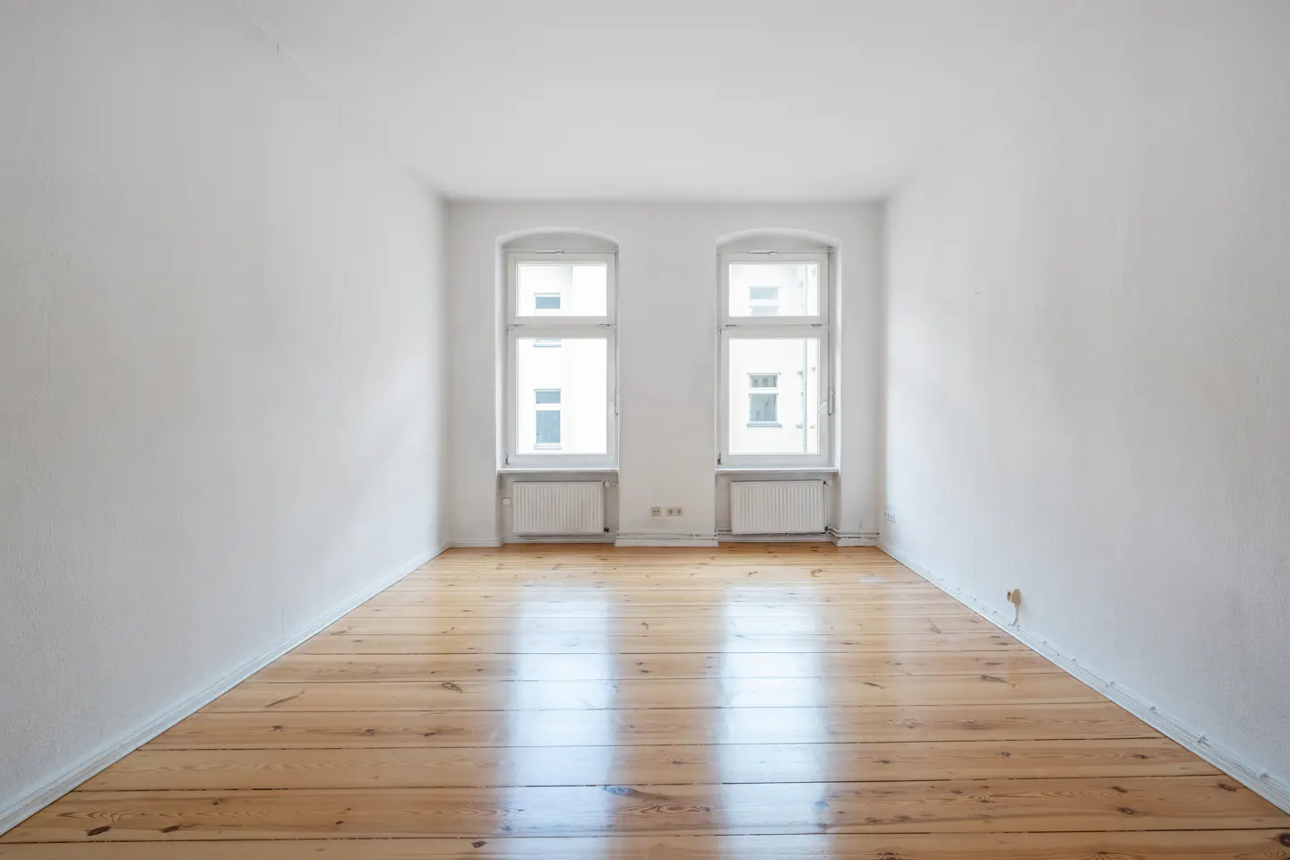 An empty room with white walls, polished light wood floors, and two windows with radiators underneath.