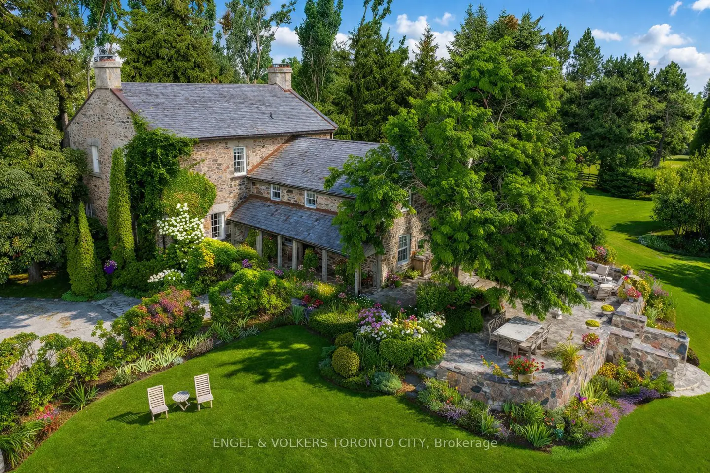 Aerial view of a stone house with a gray roof, surrounded by lush green trees, flowers, and a patio with outdoor furniture.
