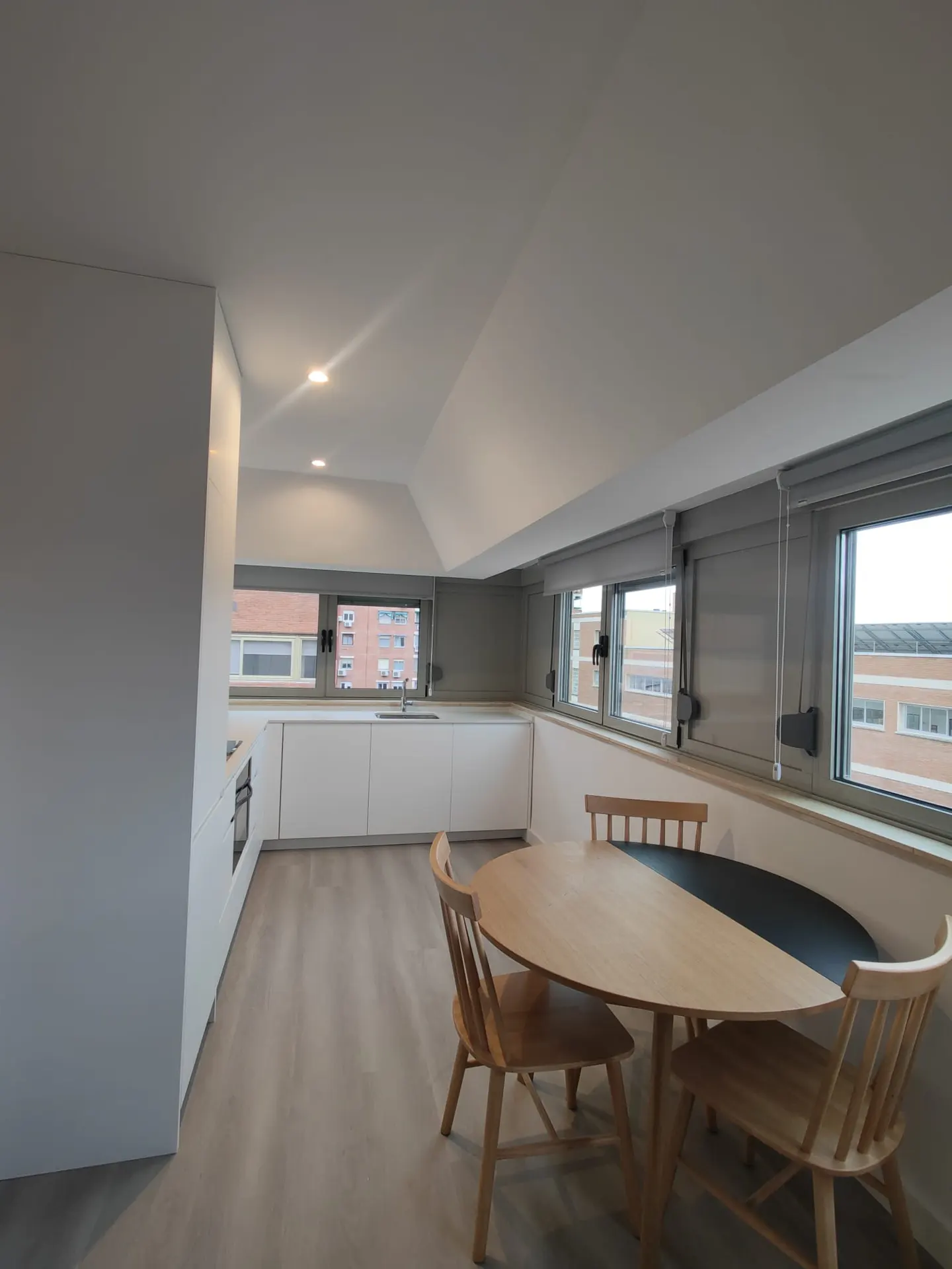 Bright, modern kitchen with white cabinets, wood floor, and a round table with four chairs. Windows line the wall above the table.