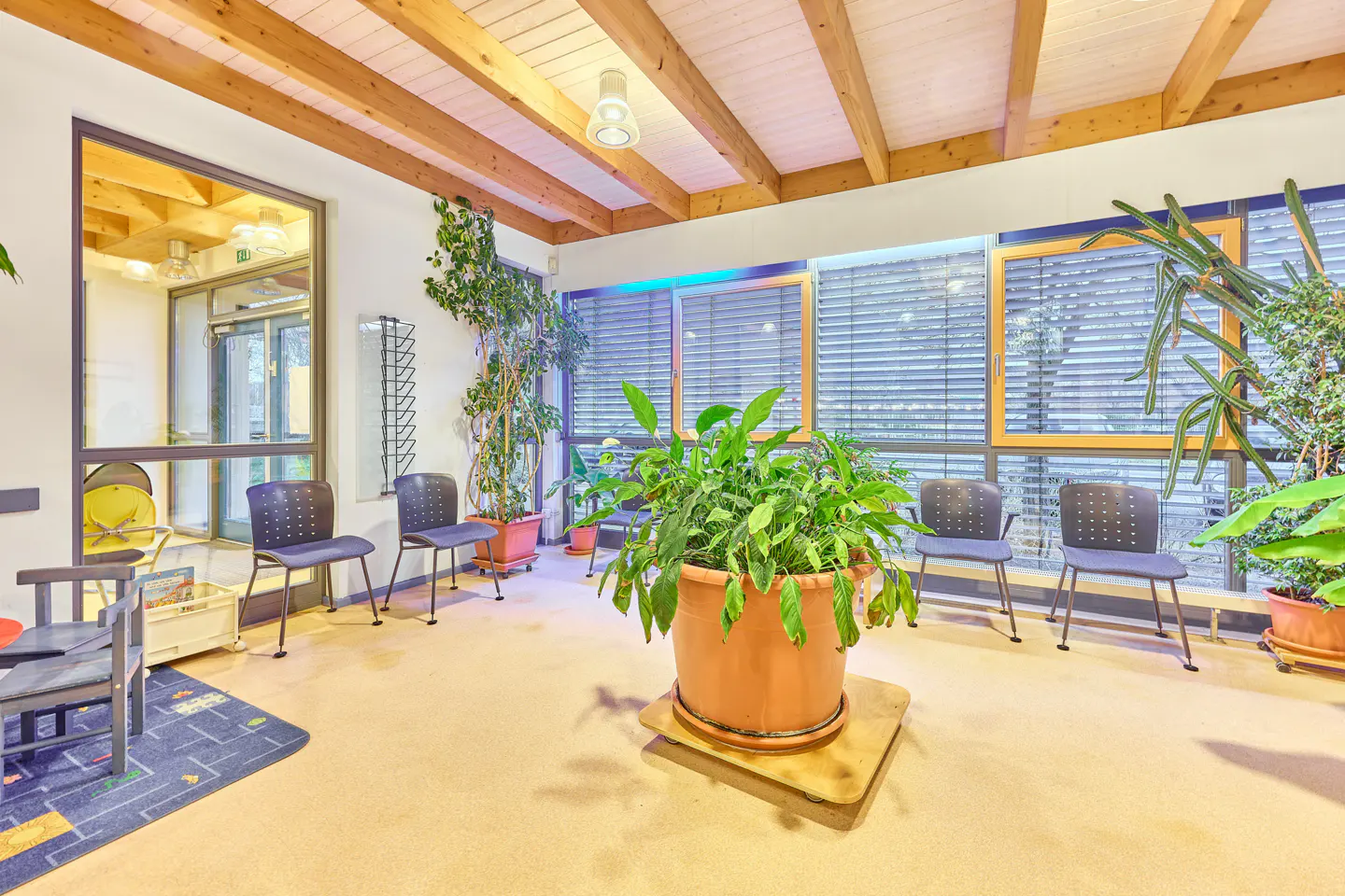 Waiting room with wooden ceiling beams, chairs, and potted plants near windows with blinds.