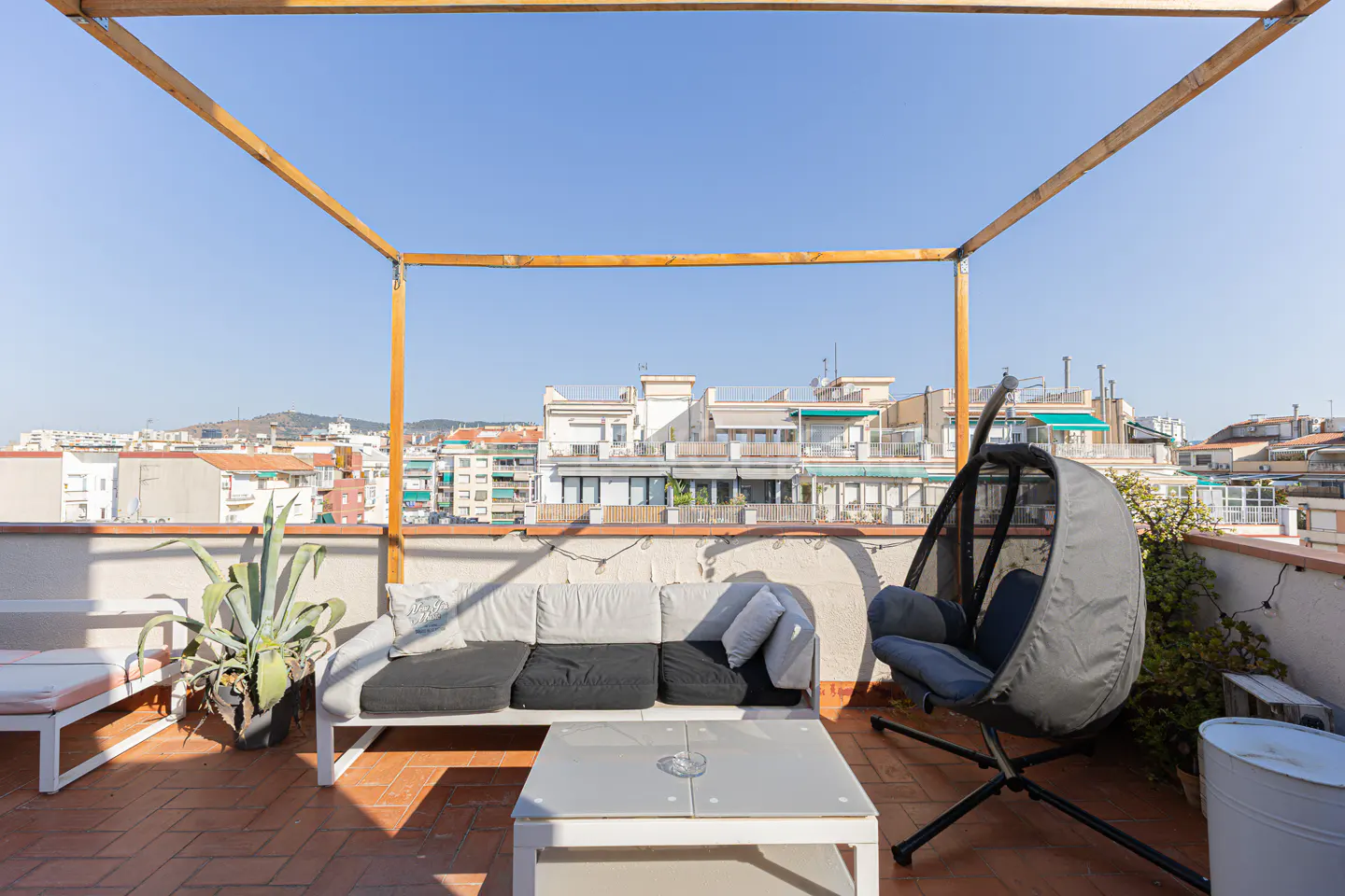 Rooftop patio with a white sofa, gray hanging chair, and white table. A wooden pergola frames the blue sky and city view.