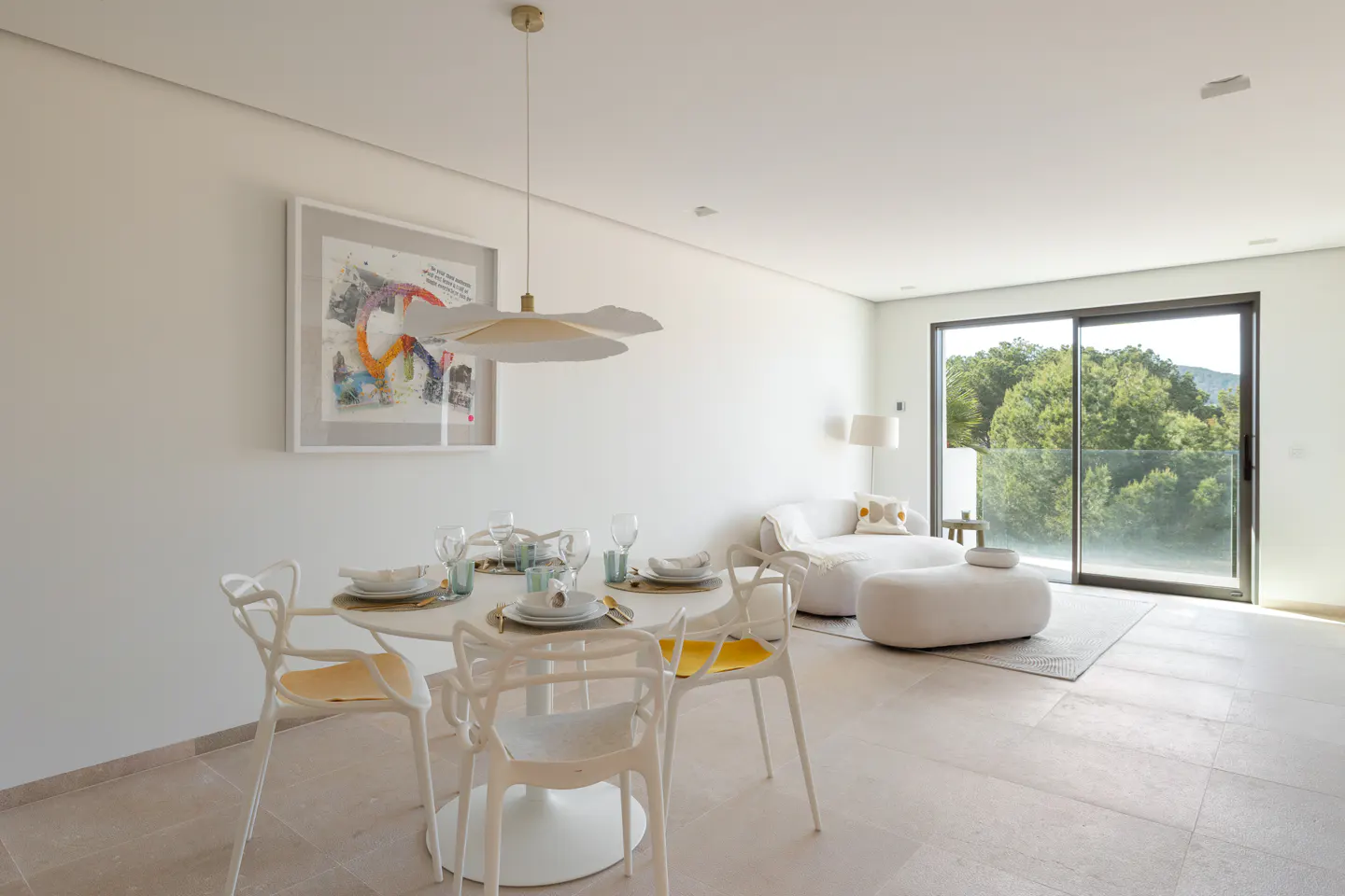 Bright, modern dining area with a round white table, white chairs with yellow cushions, and a white sofa near a sliding glass door.