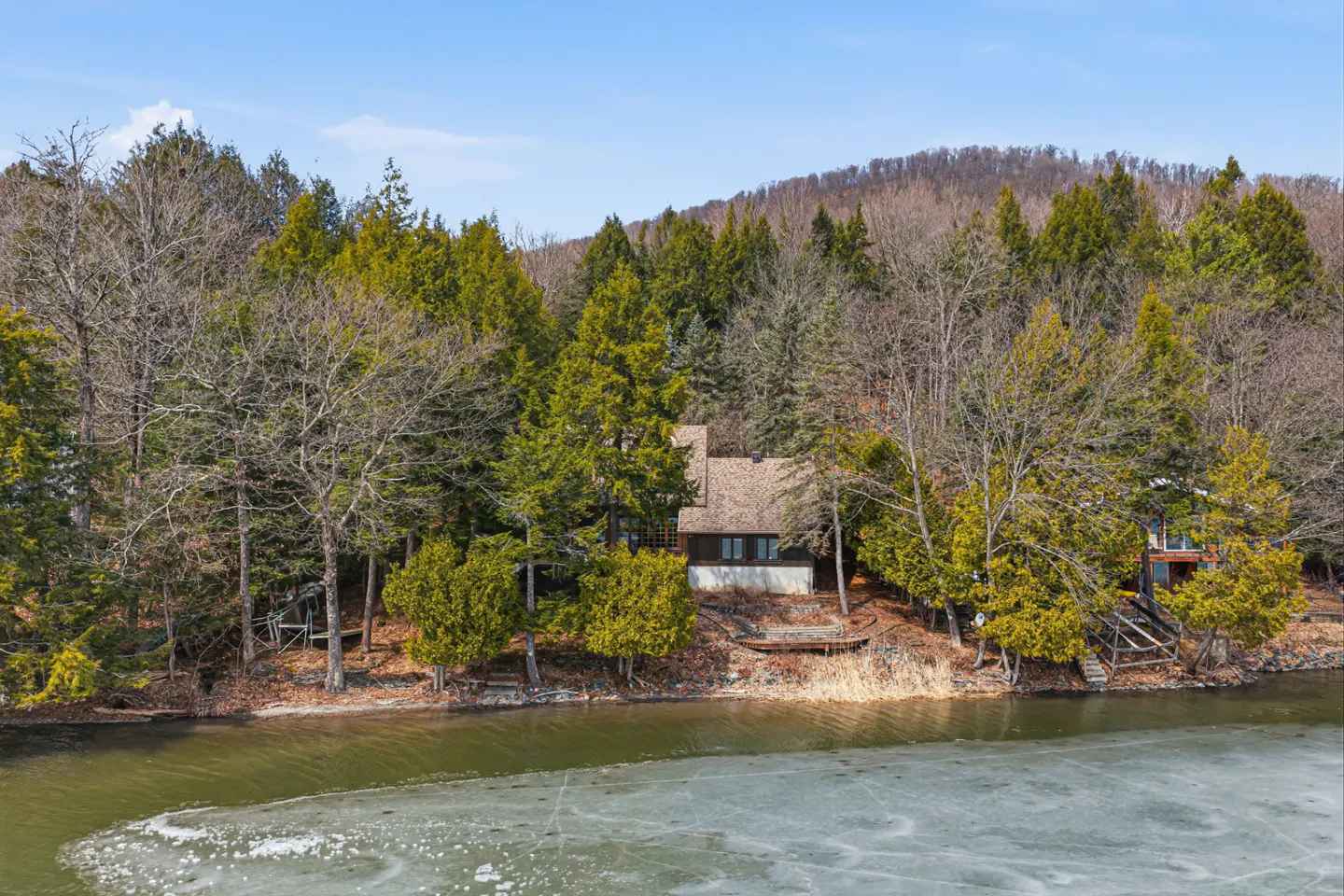 A lakefront house surrounded by trees, with a partially frozen lake in the foreground.