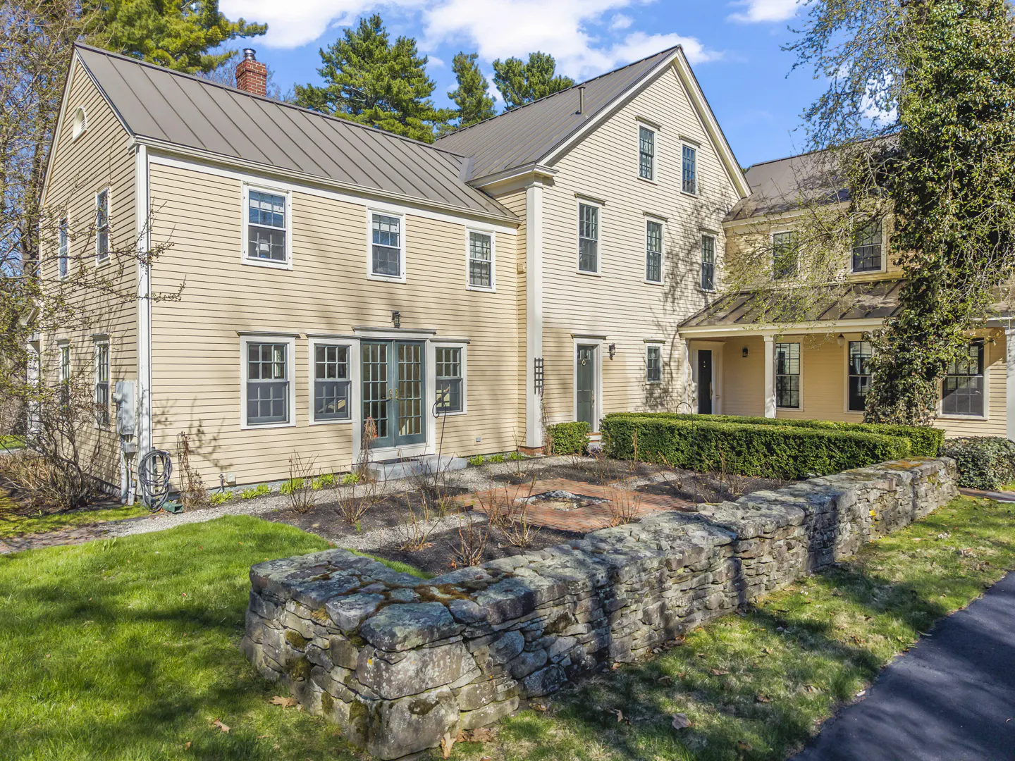 Two-story yellow house with a gray metal roof and a stone wall in the front yard.