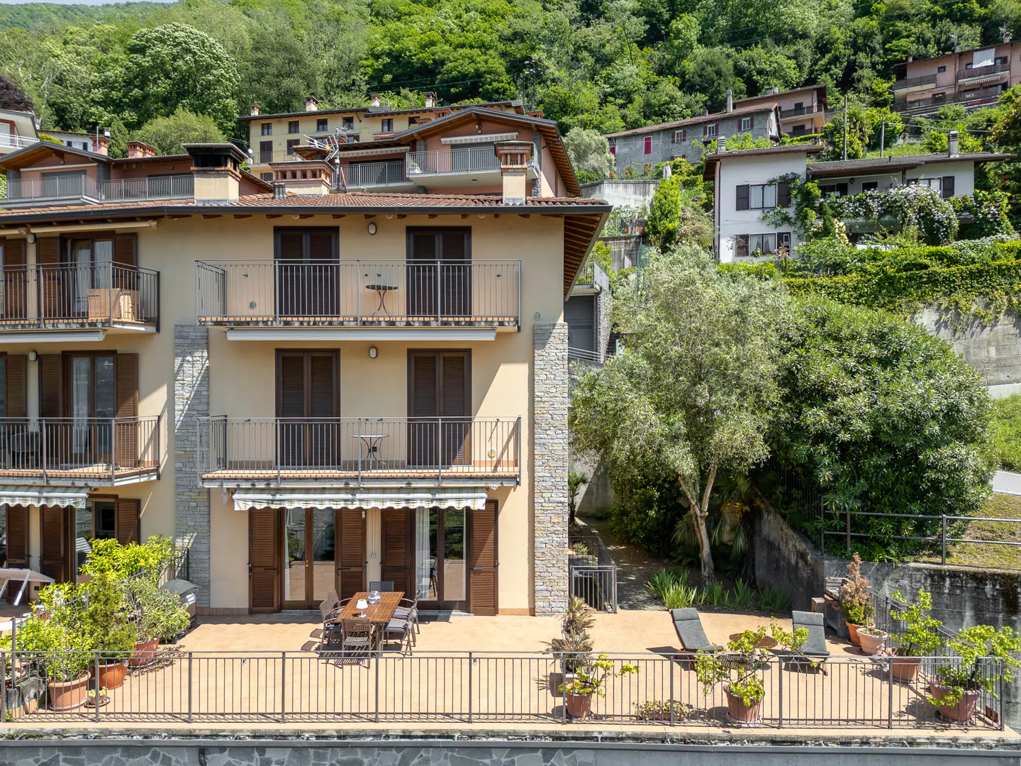 A multi-story beige apartment building with balconies and a ground-floor patio, surrounded by lush trees and hillside homes.