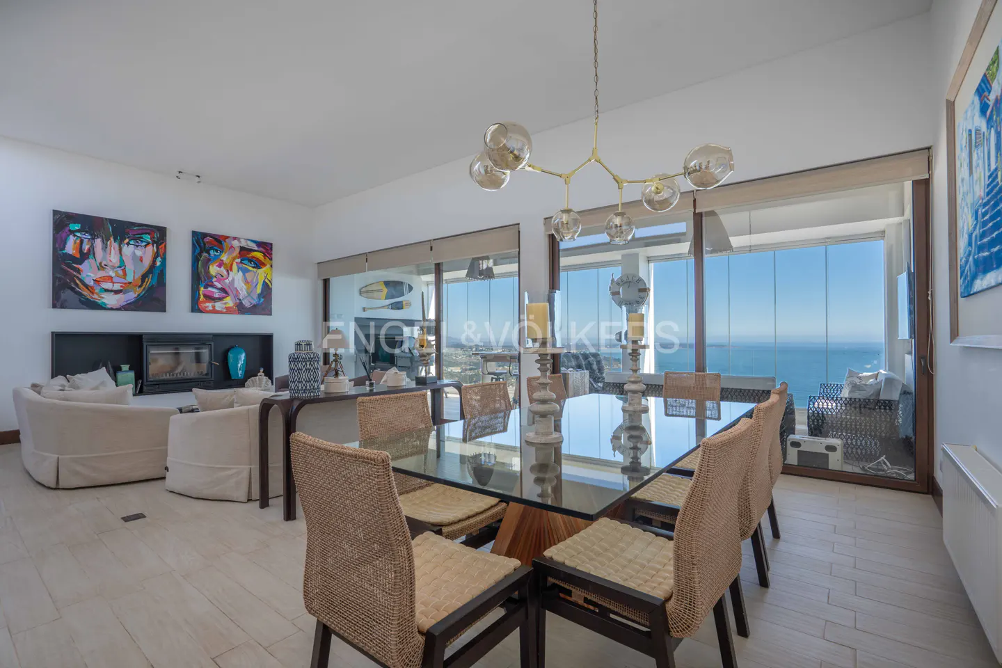 Bright, modern dining room with glass table, woven chairs, and ocean view through large windows. Art and fireplace in the background.