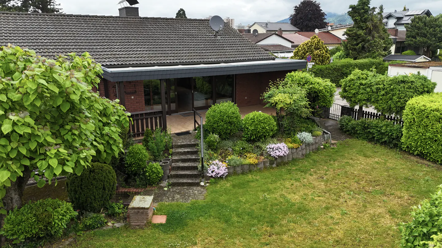 A brick house with a tiled roof, a covered patio, stone steps, and a lush garden with a lawn and trimmed hedges.