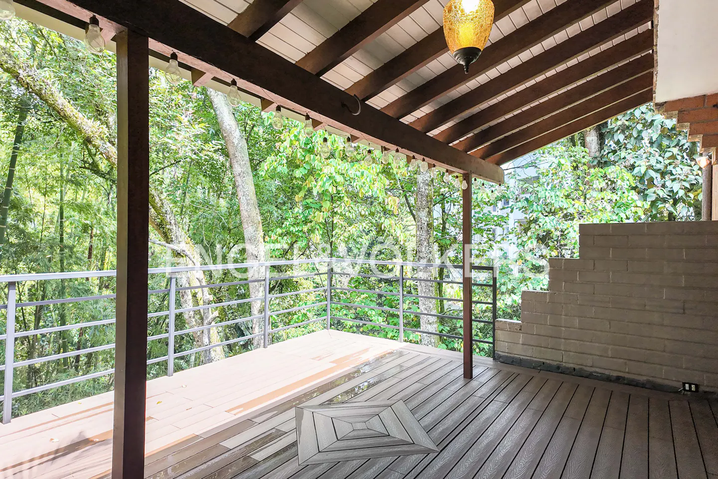 Covered wooden deck with metal railing overlooks lush green trees. Brown beams support the roof with hanging lights.