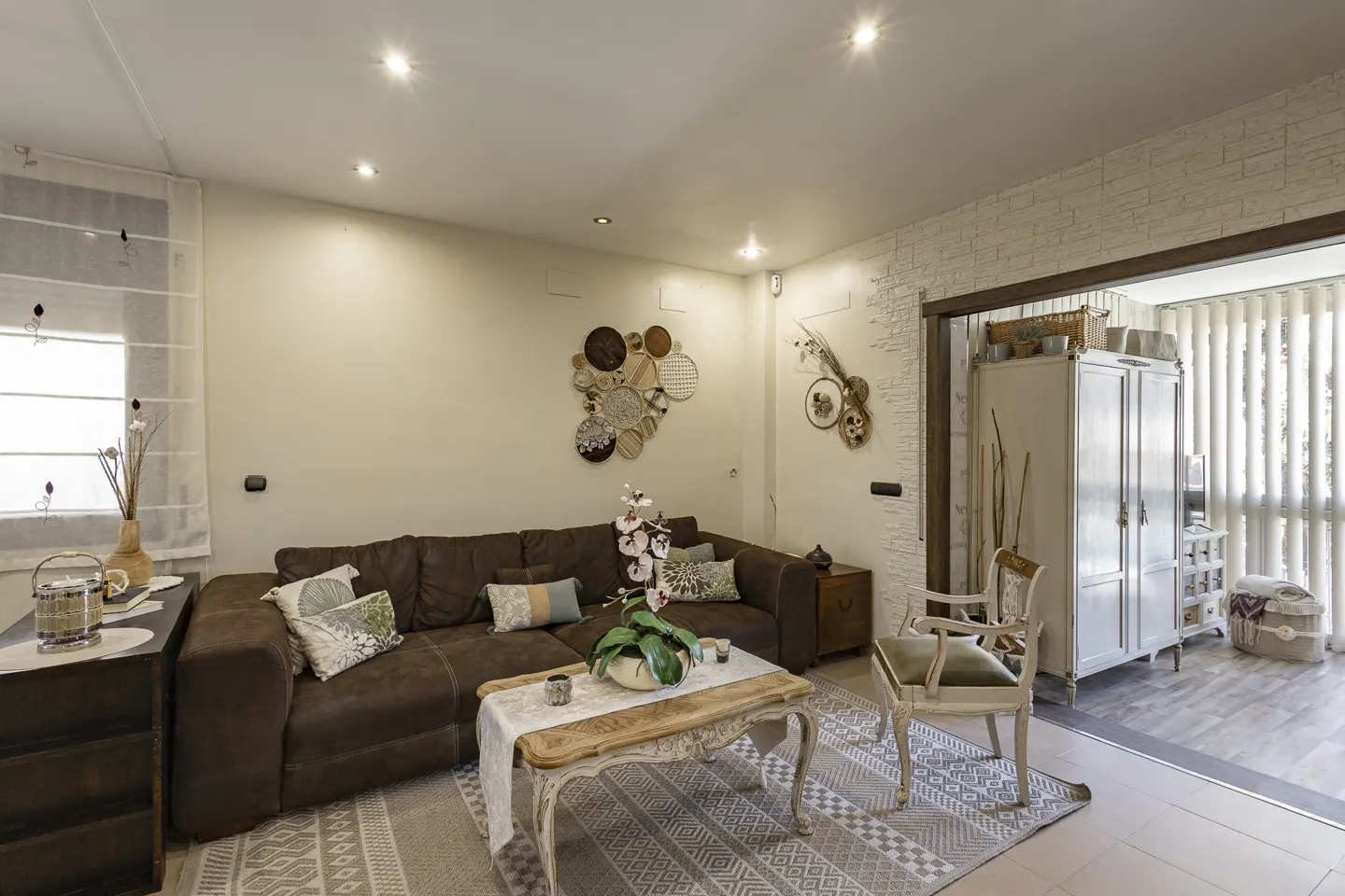 Living room with brown sectional sofa, patterned rug, and decorative wall art. A doorway leads to a bright sunroom.