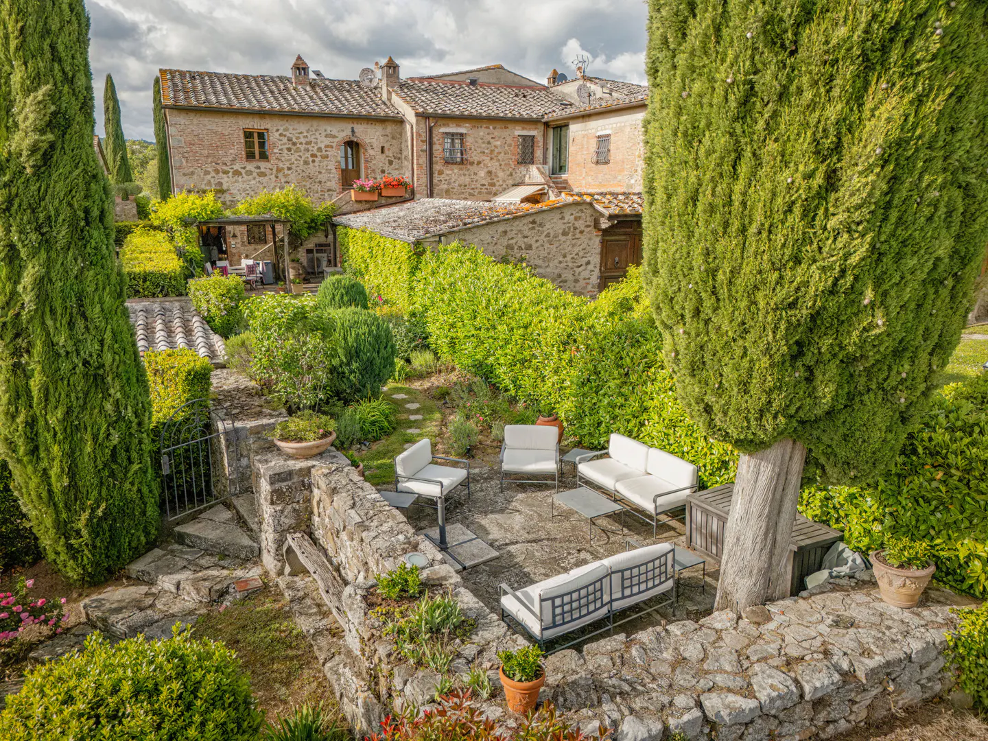 A stone patio with white outdoor furniture sits in a lush garden before a rustic stone villa in Tuscany.
