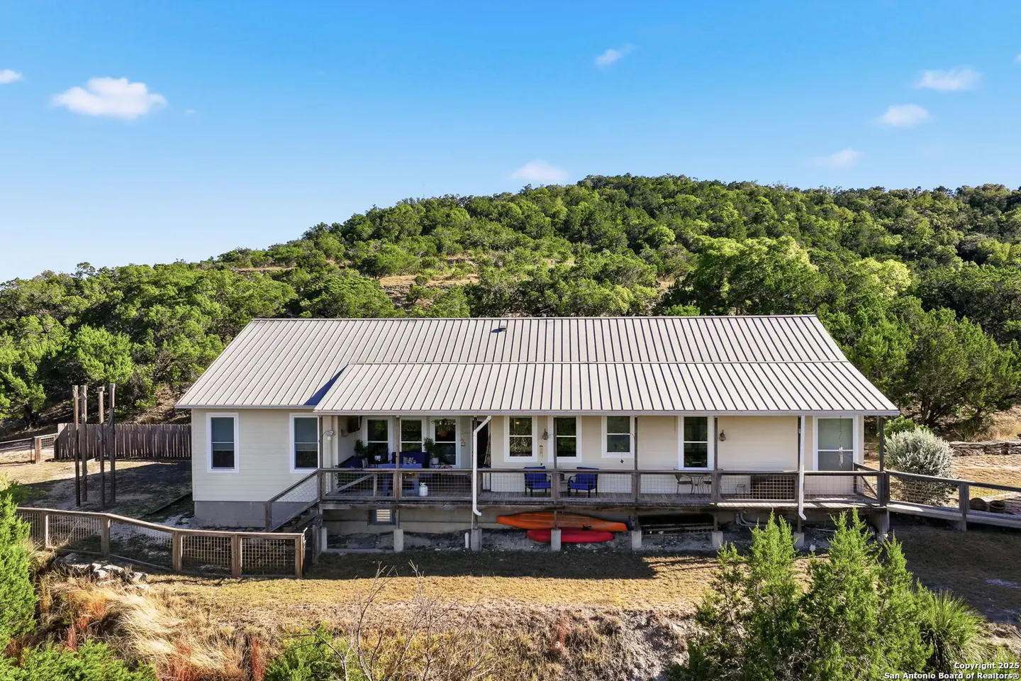 A single-story beige house with a metal roof and a wooden deck, surrounded by trees and a blue sky.