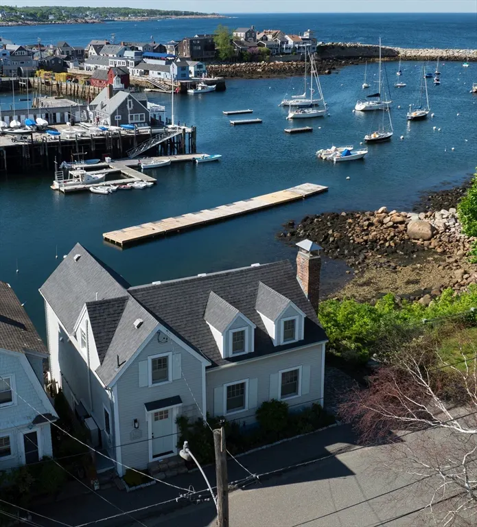 Aerial view of a light gray coastal home with a dark roof overlooking a harbor filled with sailboats and docks.