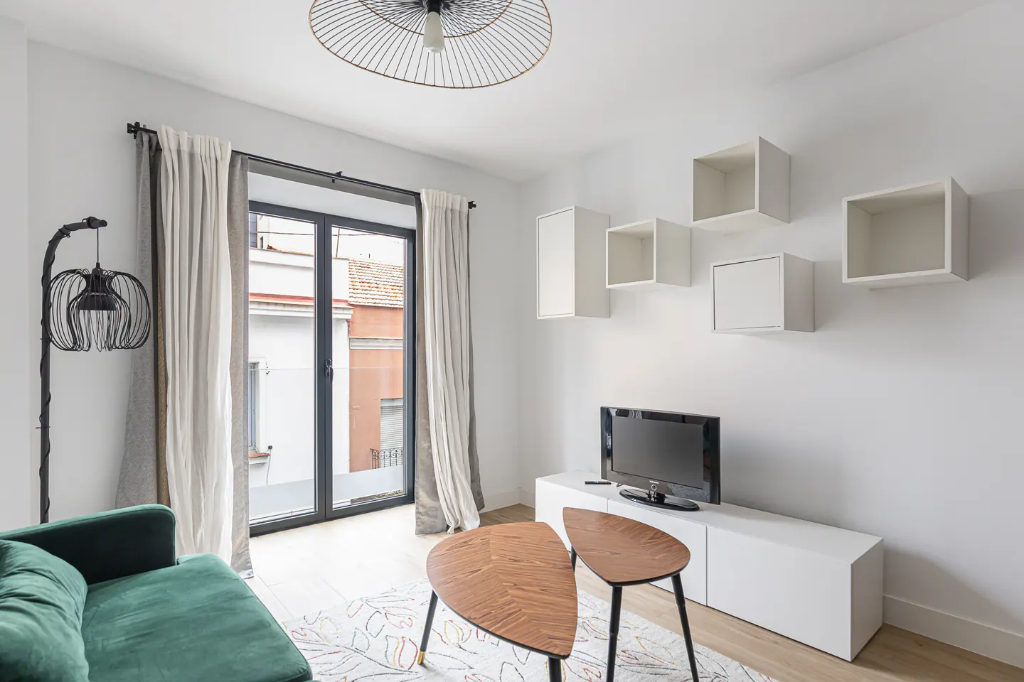Bright living room with a green sofa, wood coffee table, TV, and white shelves on a white wall. A black framed window with curtains is on the left.