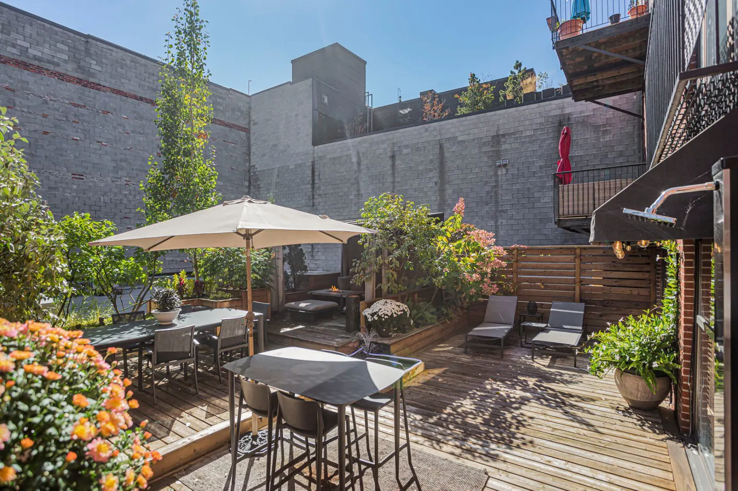 Outdoor patio with wooden deck, tables, chairs, lounge chairs, plants, and a fire pit, surrounded by brick walls.