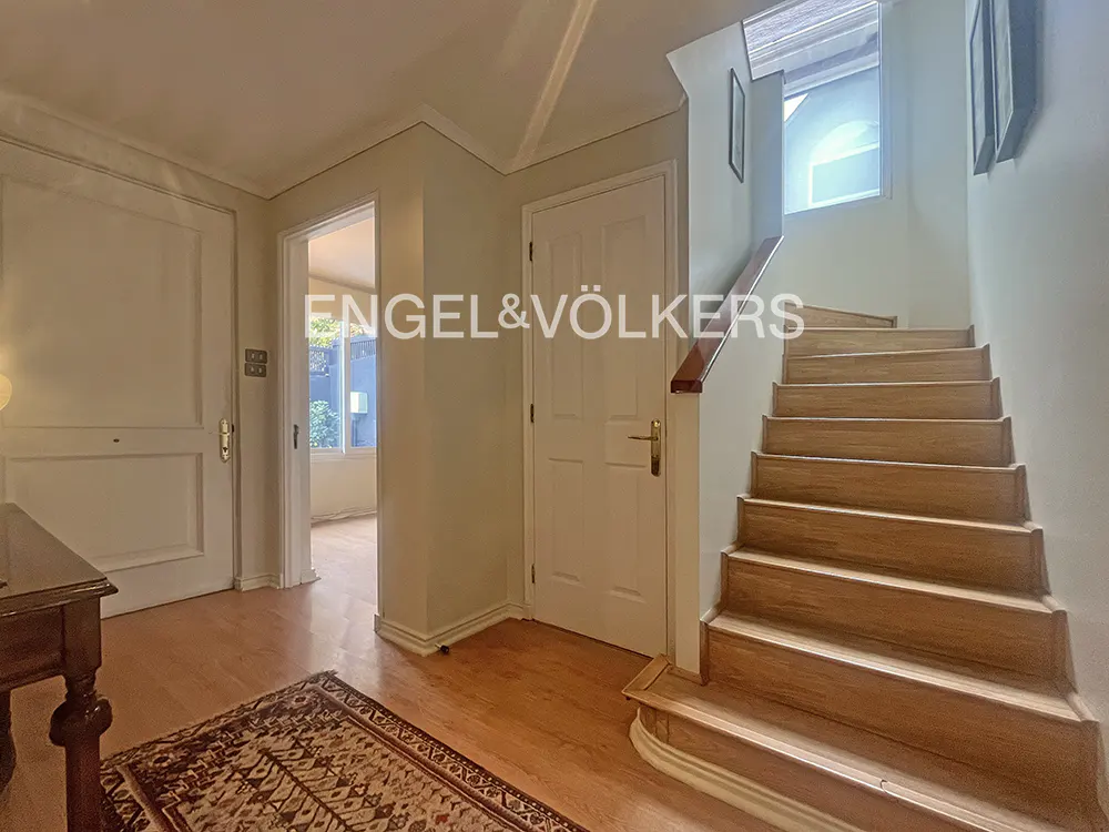 Entryway with light wood floors, white walls, and a staircase. A patterned rug sits near a dark wood table. Doors and an open doorway are visible.
