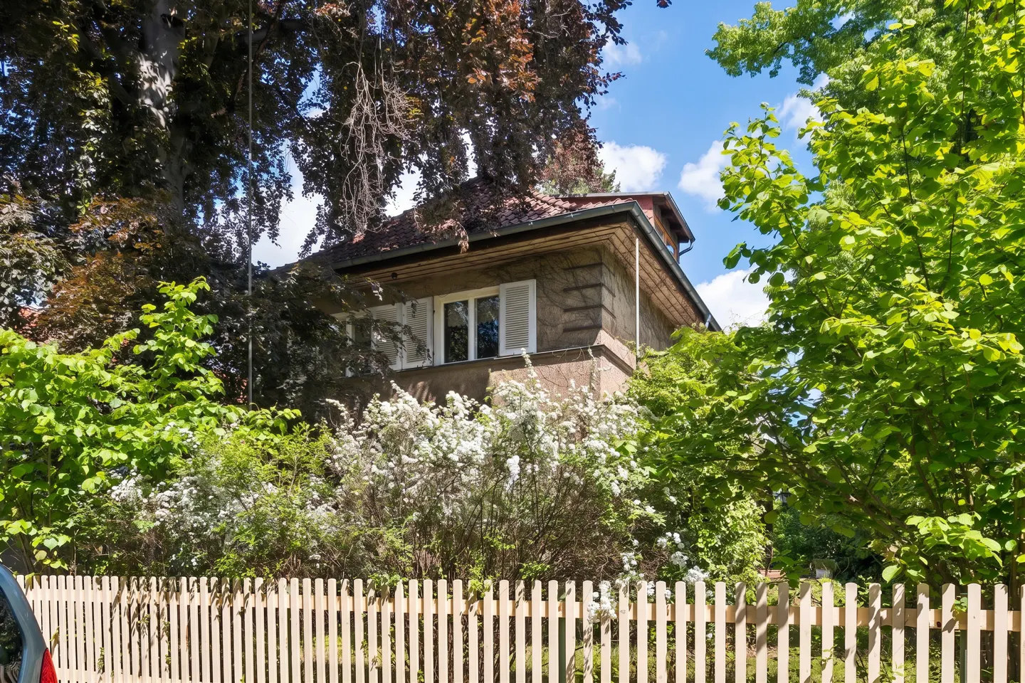 A two-story house with white shutters behind a wooden picket fence, surrounded by lush green trees and white flowers.