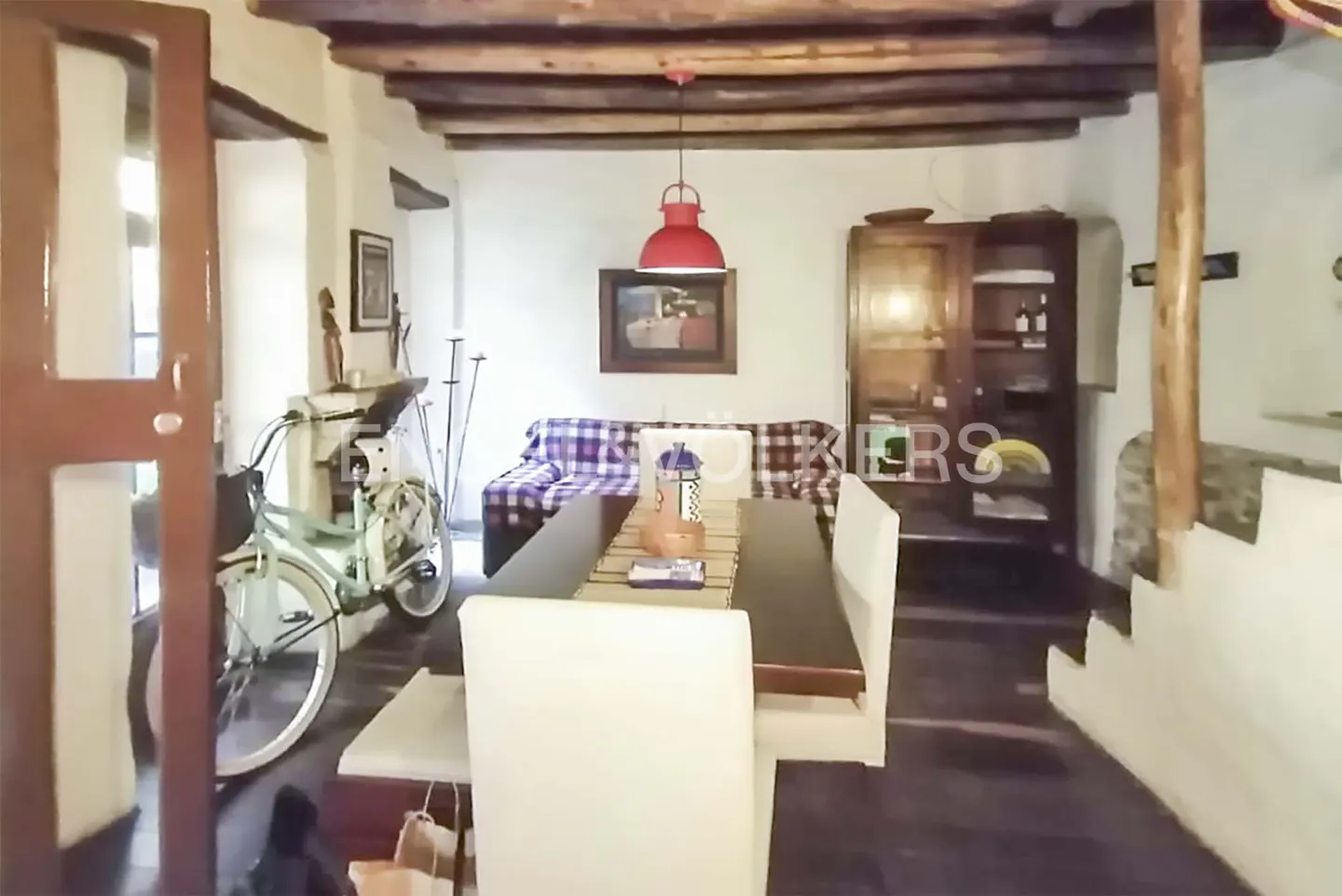 Interior view of a dining room with a wooden table, white chairs, a red pendant light, and a bicycle near the door.