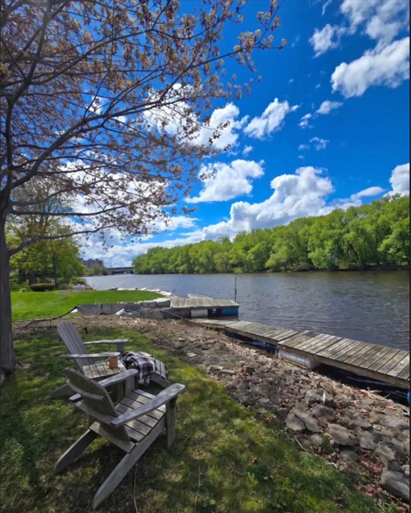 Two Adirondack chairs sit on a grassy bank near a river, with a dock, trees, and a blue sky with clouds in the background.