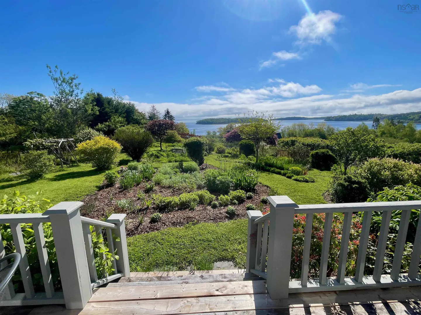 View from a wooden deck overlooking a lush garden with diverse plants, trees, and a scenic ocean bay under a blue sky.