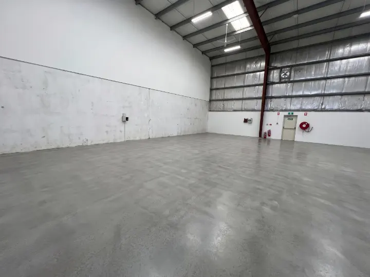 Empty warehouse interior with gray concrete floor, white walls, and a high ceiling with metal beams and fluorescent lights.