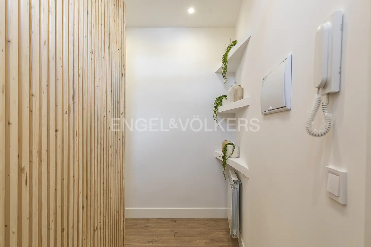Hallway with light wood slat wall, white walls, wood floor, floating shelves with plants, and intercom system.