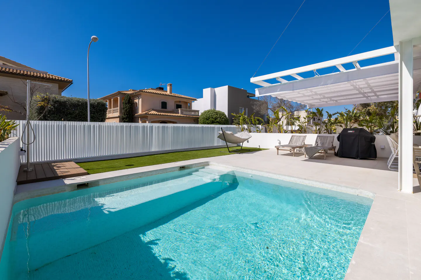 A bright, sunny backyard with a turquoise pool, white fence, and lounge chairs under a white pergola. A grill is covered nearby.