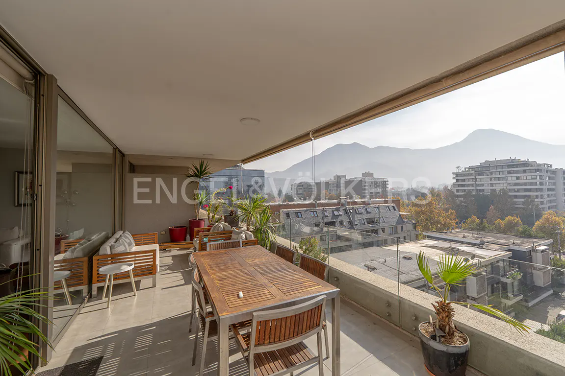 A balcony with a wooden table and chairs overlooks a city with mountains in the background.