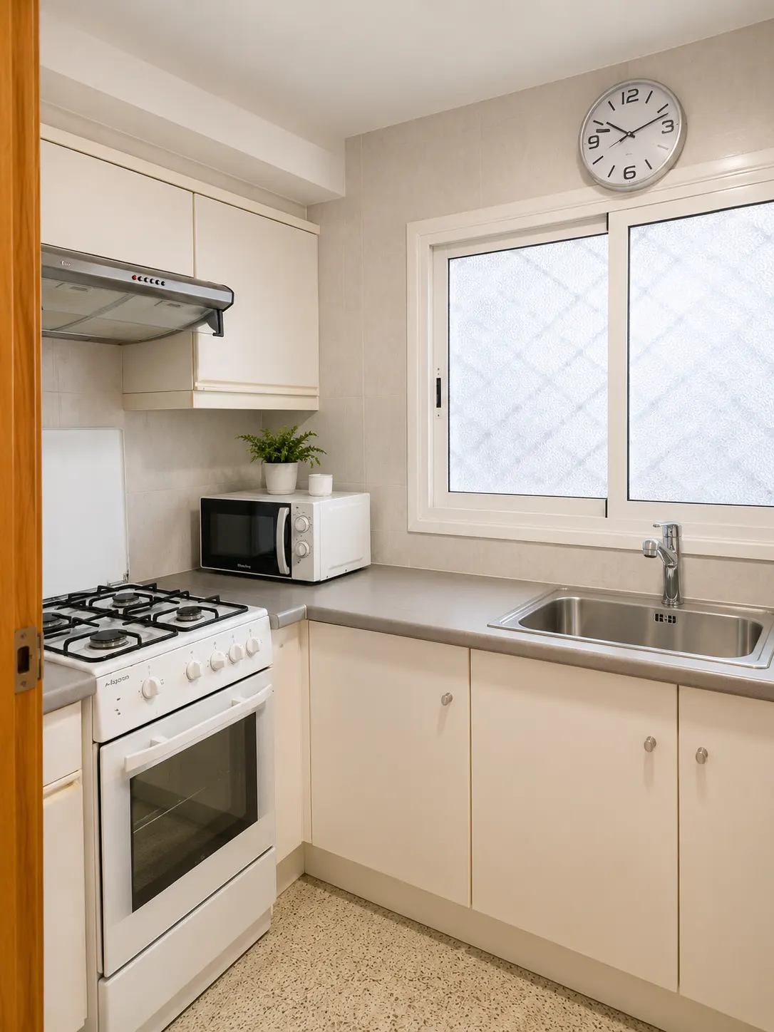 A bright kitchen with white cabinets, a white stove, a microwave, and a stainless steel sink under a window. A clock hangs on the wall.