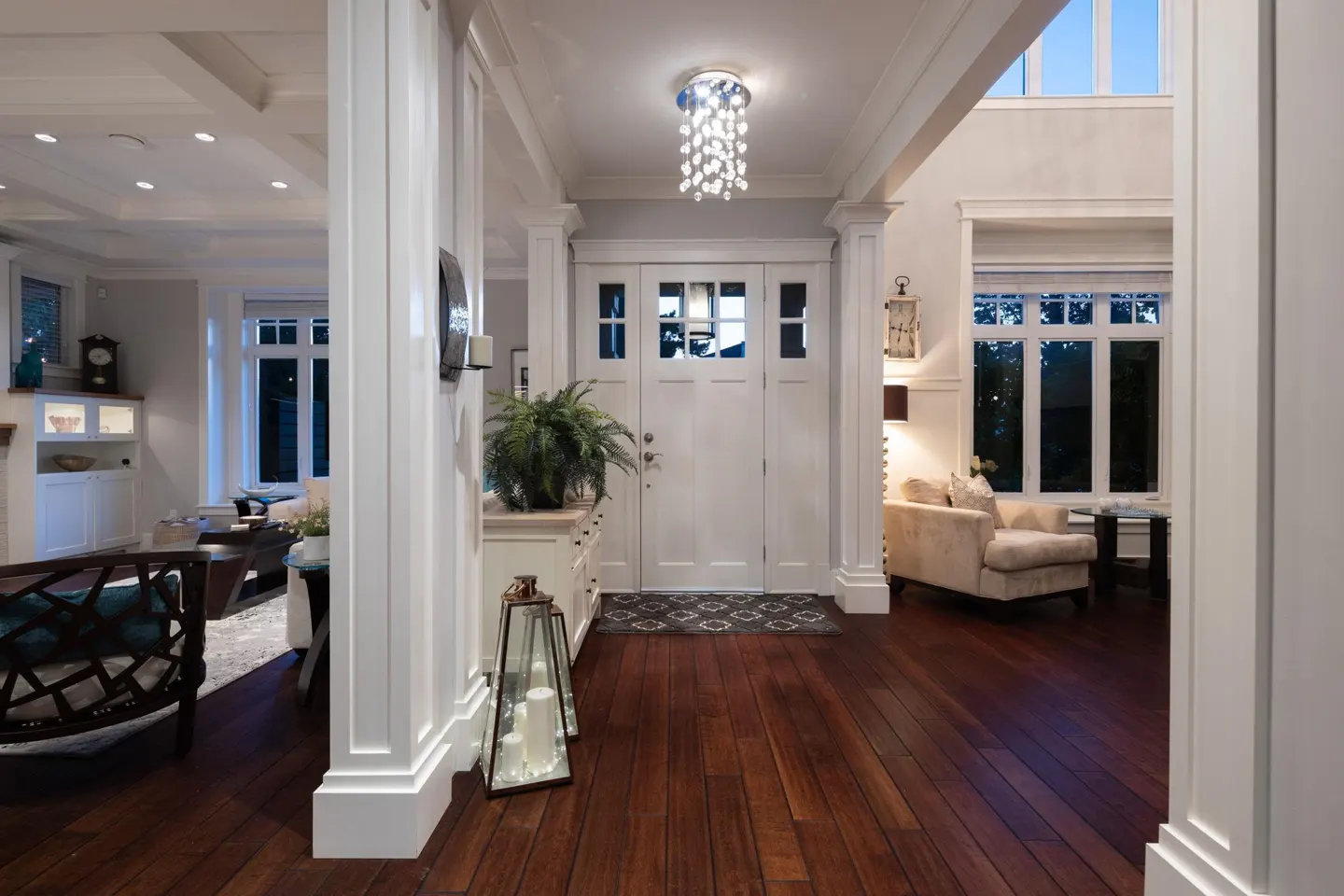 A bright foyer with dark wood floors, white trim, and a white front door with glass panes. A chandelier hangs above the door.