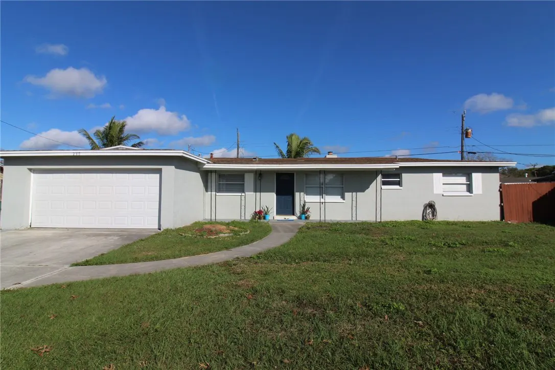 Single-story gray house with a white garage door, brown roof, and a blue front door on a green lawn under a blue sky.