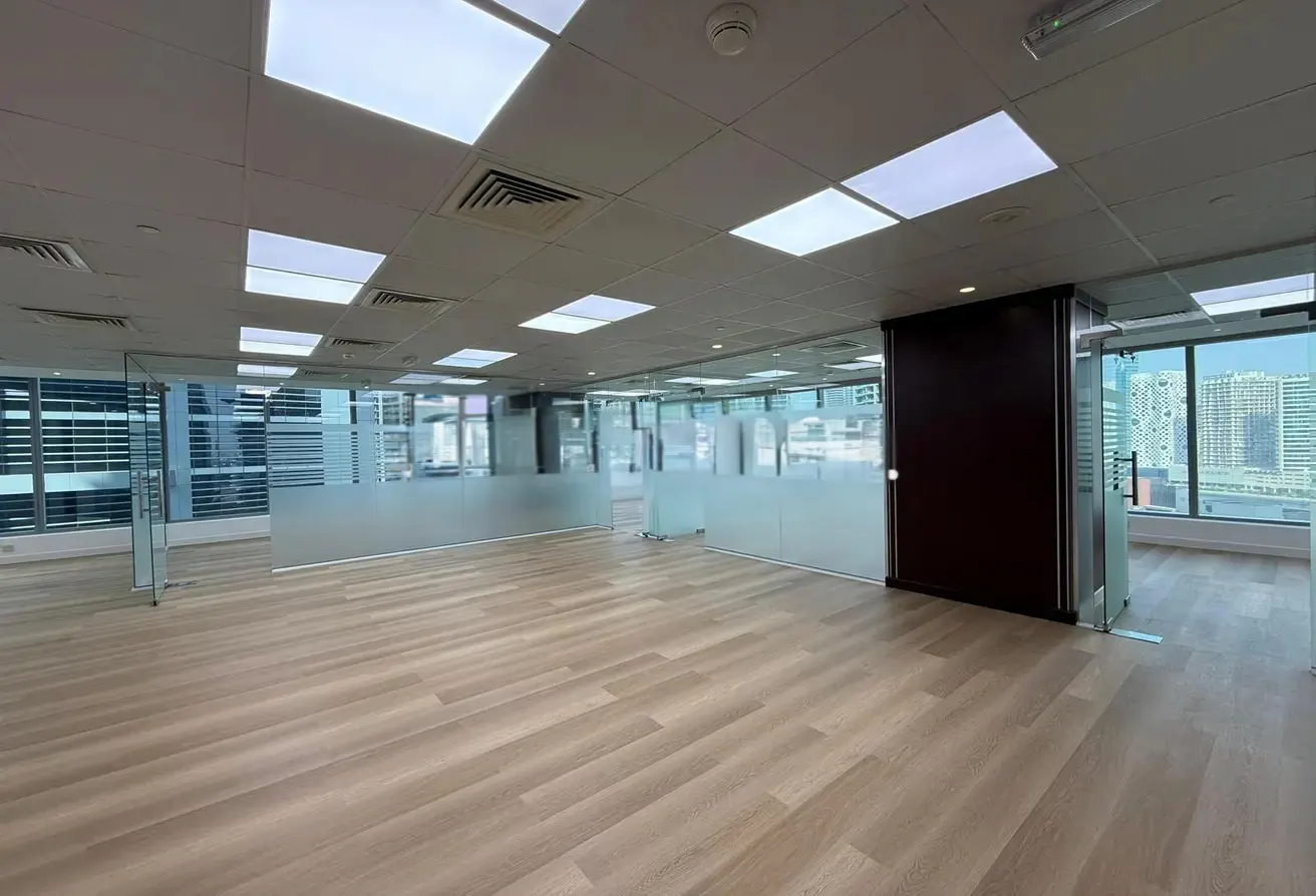 Empty office space with wood-look floors, glass walls, and a view of city buildings through large windows.