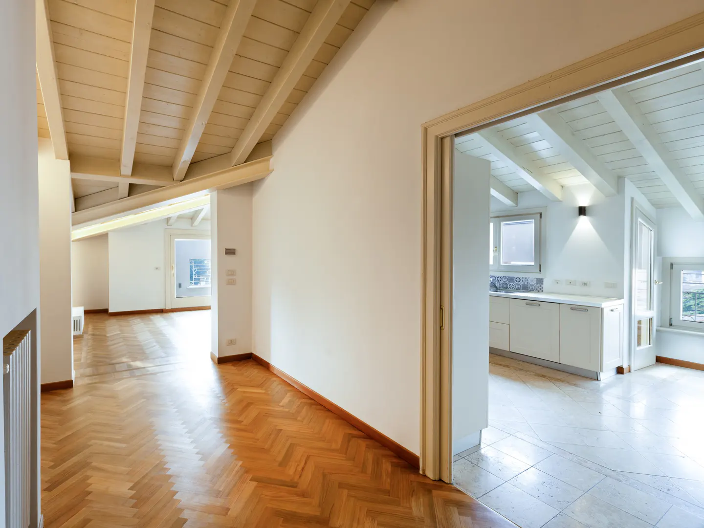 Bright, airy attic apartment with herringbone wood floors and white walls. A doorway reveals a kitchen with white cabinets and a window.
