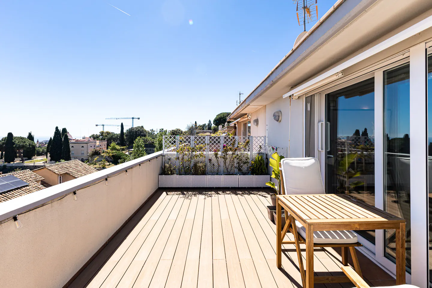 Balcony with wooden floor, table, and chair overlooking a green landscape under a clear blue sky. Sliding glass doors lead inside.