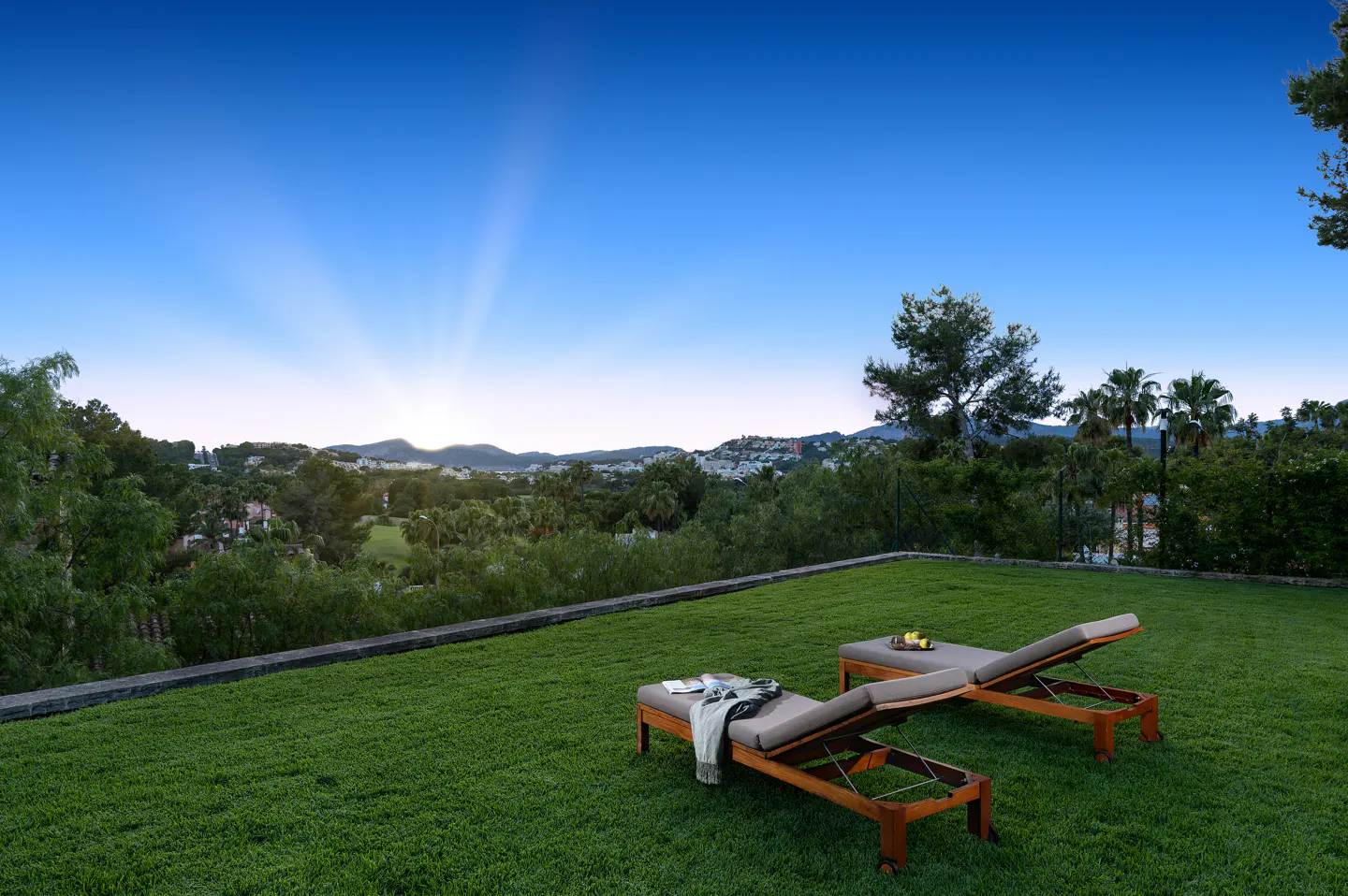 Two wooden lounge chairs on a lush green lawn overlooking a scenic valley at sunset with a clear blue sky.