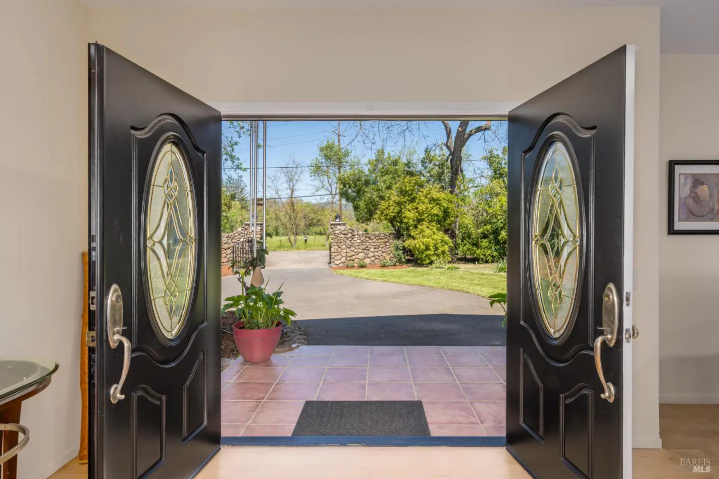 Open black double doors with oval glass panels lead to a tiled entryway and a view of a driveway and green landscape.