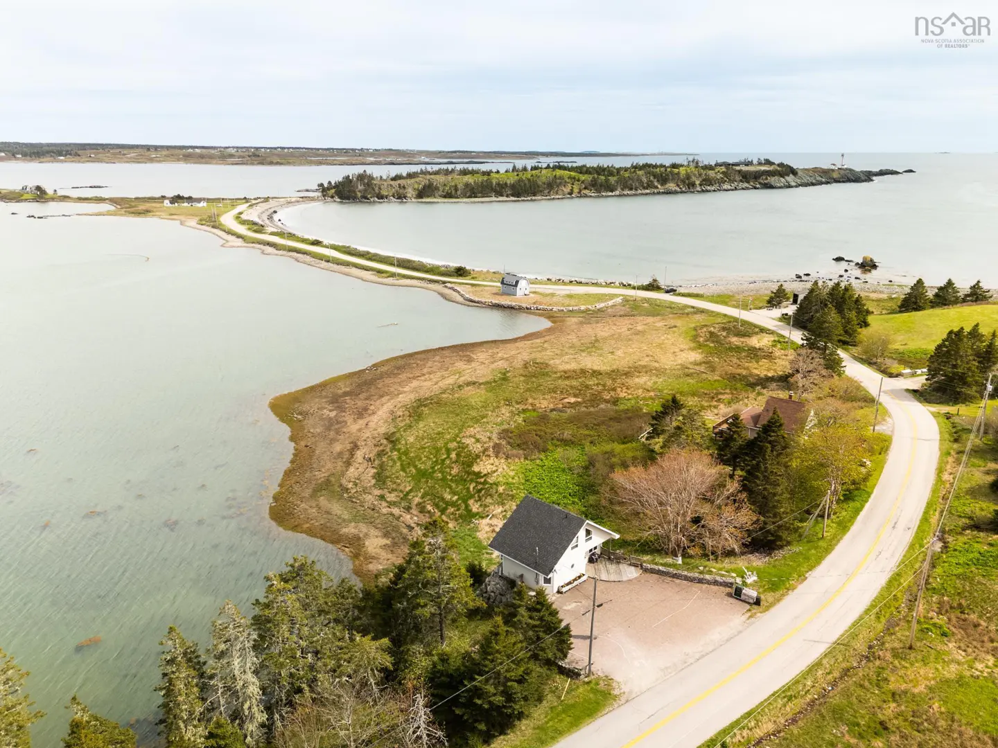 Aerial view of a coastal property with a white house, road, and ocean. Trees and green grass surround the house. A causeway leads to an island with a lighthouse.
